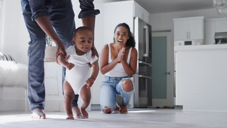 Parents-Encouraging-Smiling-Baby-Son-To-Take-First-Steps-And-Walk-At-Home