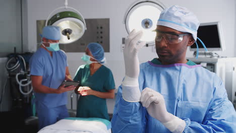 Retrato-De-Un-Cirujano-Masculino-Vistiendo-Batas-Poniéndose-Guantes-De-Látex-En-El-Quirófano-Del-Hospital