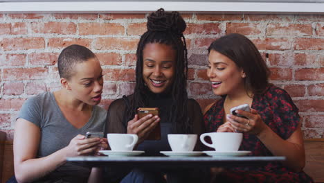 Three-Female-Friends-Meeting-For-Coffee-Sitting-At-Table-Looking-At-Mobile-Phones