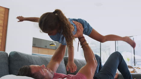 Father-Playing-Game-With-Daughter-Lying-On-Sofa-And-Lifting-Her-In-The-Air