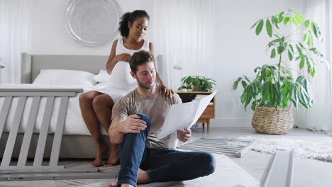 Interracial-Couple-With-Pregnant-Woman-Looking-At-Instructions-For-Building-Self-Assembly-Baby-Cot
