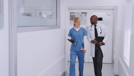 Doctor-In-White-Coat-And-Nurse-In-Scrubs-Having-Discussion-Over-Digital-Tablet-In-Hospital-Corridor