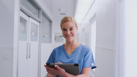 Portrait-Of-Smiling-Female-Doctor-Wearing-Scrubs-In-Hospital-Corridor-Using-Digital-Tablet