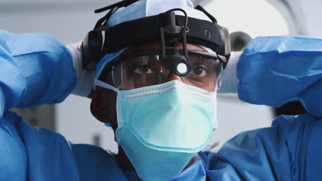 Männlicher-Chirurg-Mit-Schutzbrille-Und-Kopflicht,-Der-Im-Operationssaal-Des-Krankenhauses-Eine-Maske-Aufsetzt