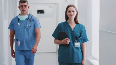 Portrait-Of-Smiling-Female-Doctor-Wearing-Scrubs-In-Busy-Hospital-Corridor-Holding-Digital-Tablet