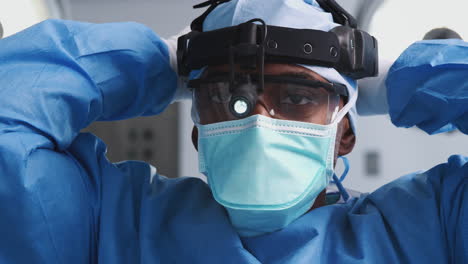 Cirujano-Masculino-Con-Gafas-Protectoras-Y-Luz-De-Cabeza-Poniéndose-Una-Máscara-En-El-Quirófano-Del-Hospital