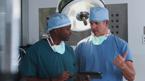 Two-Male-Surgeons-Wearing-Scrubs-Looking-At-Digital-Tablet-In-Hospital-Operating-Theater