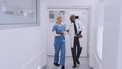 Médico-Con-Bata-Blanca-Y-Enfermera-Con-Uniforme-Médico-Discutiendo-Sobre-Una-Tableta-Digital-En-El-Pasillo-Del-Hospital