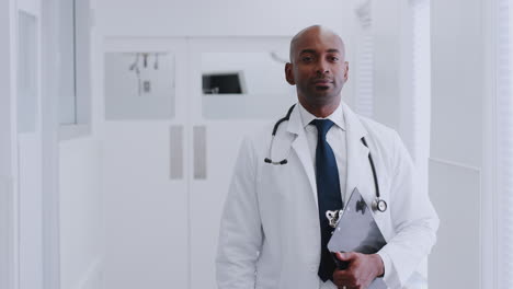 Portrait-Of-Mature-Male-Doctor-Wearing-White-Coat-Standing-In-Hospital-Corridor-Holding-Clipboard