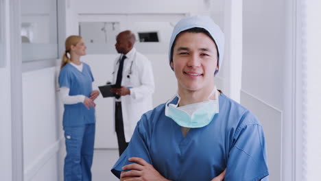 Male-Surgeon-Wearing-Scrubs-And-Mask-Standing-In-Hospital-Corridor-With-Colleagues-In-Background
