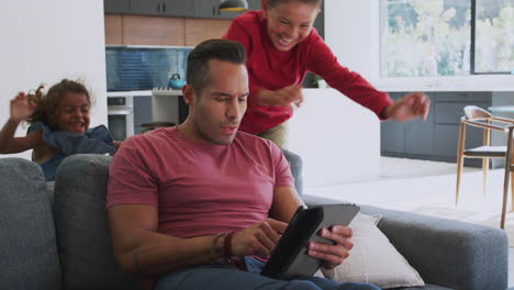 Children-Creeping-Up-And-Surprising-Father-Using-Digital-Tablet-At-Home