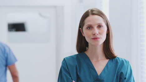 Head-And-Shoulders-Portrait-Of-Female-Doctor-Wearing-Scrubs-In-Busy-Hospital-Corridor