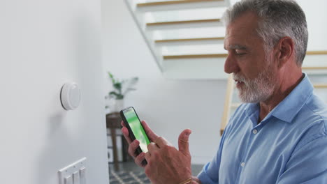 Älterer-Hispanischer-Mann-Nutzt-App-Am-Telefon,-Um-Den-Digitalen-Zentralheizungsthermostat-Zu-Hause-Zu-Steuern