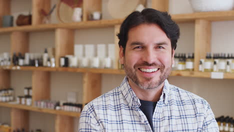 Portrait-Of-Male-Owner-Of-Gift-Store-Standing-In-Front-Of-Shelves-With-Cosmetics-And-Candles