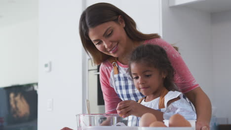 Hispanic-Mother-And-Daughter-Cracking-Eggs-Into-Bowl-As-They-Bake-Cake-In-Kitchen