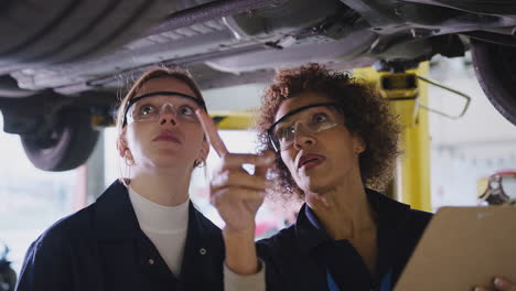 Female-Tutor-With-Student-Looking-Underneath-Car-On-Hydraulic-Ramp-On-Auto-Mechanic-Course