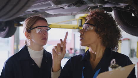 Female-Tutor-With-Student-Looking-Underneath-Car-On-Hydraulic-Ramp-On-Auto-Mechanic-Course