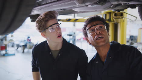 Male-Tutor-With-Student-Looking-Underneath-Car-On-Hydraulic-Ramp-On-Auto-Mechanic-Course