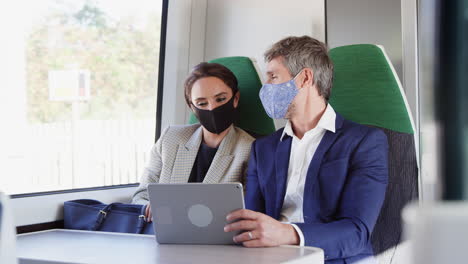Businesswoman-And-Businessman-On-Train-Using-Digital-Tablet-Wearing-PPE-Face-Mask-During-Pandemic