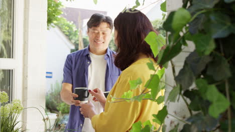 Mature-Asian-woman-planting-wooden-container-in-summer-garden-as-husband-brings-her-hot-drink-in-cup---shot-in-slow-motion