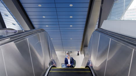 Businessman-On-Escalator-At-Railway-Station-With-Mobile-Phone-Wearing-PPE-Face-Mask-During-Pandemic