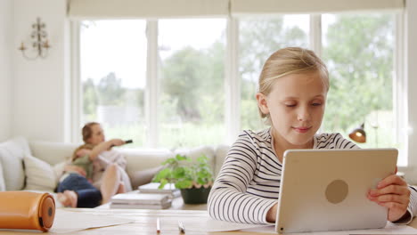 Girl-At-Table-With-Digital-Tablet-Home-Schooling-During-Health-Pandemic-With-Family-In-Background