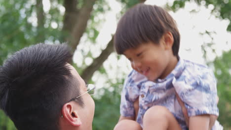 Asiatischer-Vater-Hebt-Lachenden-Sohn-In-Die-Luft,-Während-Sie-Gemeinsam-Im-Garten-Spielen