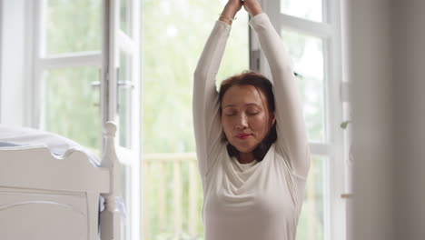 Mature-Asian-woman-in-pyjamas-sitting-on-bedroom-floor-meditating-in-yoga-pose---shot-in-slow-motion