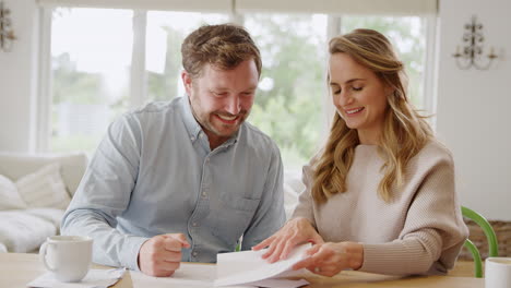 Excited-Couple-Sitting-At-Table-At-Home-Opening-Letter-With-Good-Financial-News