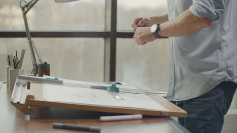 architect-man-Standing-working-with-blueprints-sketching-a-construction-project-on-wood-desk-at-home-office.Construction-design-concept.vintage-color-tone.-Engineer-works-in-a-bright-office-with-a-large-window-concentrates-and-draws-blueprints.-Workplace-of-an-architect-or-designer:-loft-style-minim
