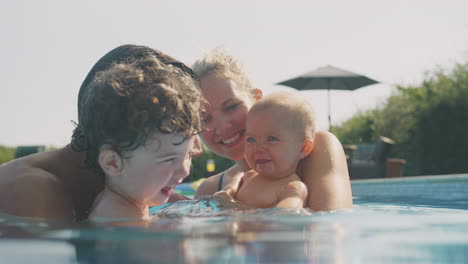 Family-With-Young-Son-And-Baby-Having-Fun-On-Summer-Vacation-Playing-And-Splashing-In-Swimming-Pool