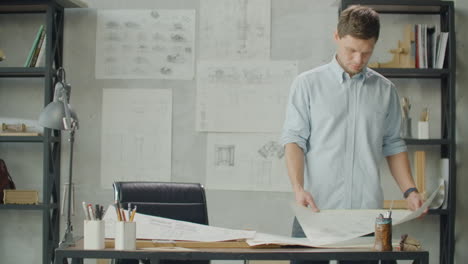 Bearded-man-hipster-standing-faces-to-panoramic-window-and-looks-at-blueprints.-Young-architect-in-stylish-loft-office-works-with-documents.-Creative-architect-thinking-on-the-big-drawings.
