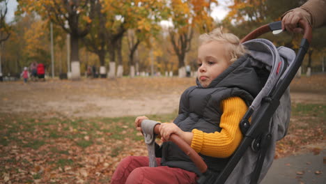 portrait-of-little-child-in-stroller-mother-is-rolling-his-in-park-area-in-city-at-autumn-day-rest-and-walk-at-weekend
