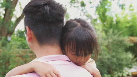 Liebevoller-Asiatischer-Vater-Kuschelt-Tochter-Im-Garten,-Während-Das-Mädchen-über-Seine-Schulter-Schaut