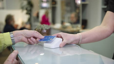 Close-Up-Of-Senior-Woman-Making-Contactless-Payment-To-Stylist-In-Salon-With-Credit-Card