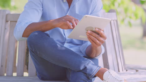 Close-Up-Of-Man-Sitting-On-Bench-Under-Tree-In-Summer-Park-Using-Digital-Tablet