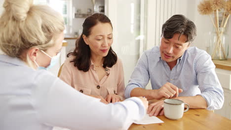 Mature-Asian-couple-at-home-meeting-with-female-financial-advisor-in-mask-during-pandemic-and-signing-document--shot-in-slow-motion