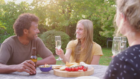 Group-Of-Friends-Celebrating-With-Beer-And-Champagne-As-They-Sit-At-Table-In-Garden-With-Snacks