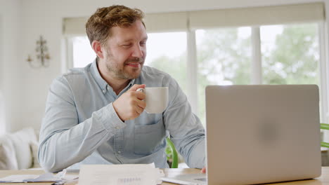 Smiling-Man-With-Laptop-Sitting-At-Table-At-Home-Reviewing-Domestic-Finances