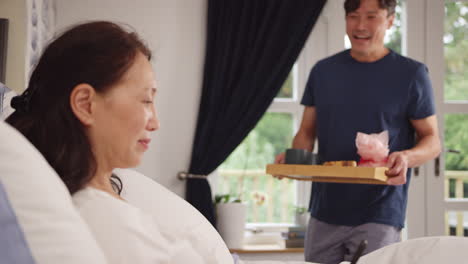 Mature-Asian-man-bringing-wife-using-mobile-phone-breakfast-in-bed-to-celebrate-birthday-or-anniversary---shot-in-slow-motion