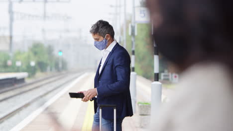 Business-Commuters-On-Railway-Platform-With-Mobile-Phones-Wearing-PPE-Face-Masks-During-Pandemic