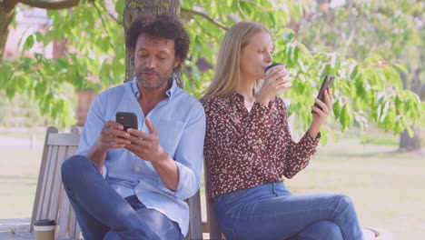 Mature-Couple-Meeting-In-Outdoor-Park-Sitting-On-Bench-Looking-At-Mobile-Phones-Together