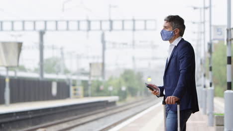 Geschäftsmann-Auf-Bahnsteig-Mit-Mobiltelefon-Und-PSA-Gesichtsmaske-Während-Einer-Gesundheitspandemie