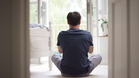 Rear-view-of-mature-Asian-man-in-pyjamas-sitting-on-bedroom-floor-meditating-in-yoga-pose---shot-in-slow-motion