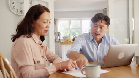 Mature-Asian-couple-sitting-at-table-at-home-using-laptop-to-organise-household-bills-and-finances---shot-in-slow-motion