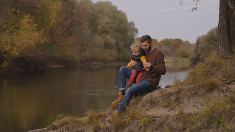 father-and-little-son-are-spending-time-together-at-shore-of-small-forest-river-resting-at-nature-at-autumn-happy-parenthood-and-childhood-memories-about-family-trip