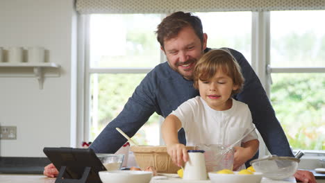 Father-And-Son-In-Pyjamas-Baking-In-Kitchen-At-Home-Following-Recipe-On-Digital-Tablet