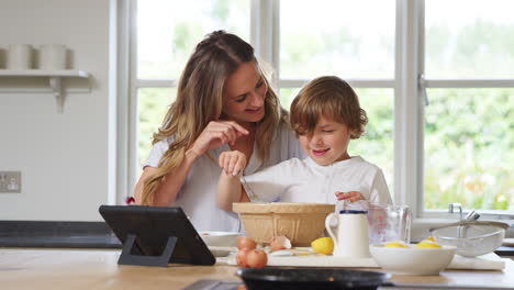 Mother-And-Son-In-Pyjamas-Baking-In-Kitchen-At-Home-Following-Recipe-On-Digital-Tablet