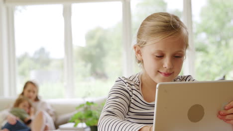 Girl-At-Table-With-Digital-Tablet-Home-Schooling-During-Health-Pandemic-With-Family-In-Background