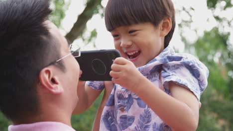 Asiatischer-Vater-Spielt-Mit-Lächelndem-Sohn-Im-Garten,-Während-Der-Junge-Sein-Mobiltelefon-In-Der-Hand-Hält